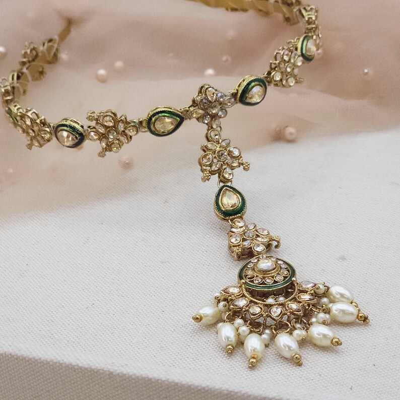 May include: Gold-toned headpiece with intricate floral and teardrop designs. The headpiece features clear gemstones and small white pearls. Green enamel accents the design. The headpiece is likely for a special occasion.