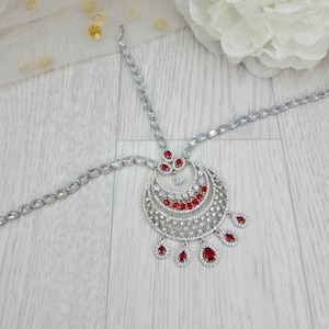 Może przedstawiać: Srebrna i czerwona ozdobna opaska na głowę z motywem półksiężyca, ozdobiona kamieniami szlachetnymi. Opaska ma łańcuszek, który owija się wokół głowy, a w środku zwisa wisiorek.