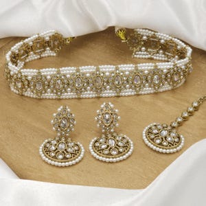 Puede incluir: Un conjunto de collar gargantilla de oro y perlas blancas con pendientes y tikka a juego. El collar presenta un diseño delicado con piedras transparentes y perlas.