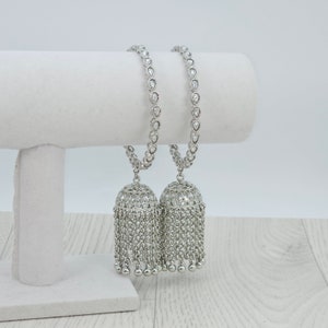 May include: A pair of silver bangles with dangling charms. The bangles are adorned with clear stones and the charms are shaped like bells with a fringe of silver beads.