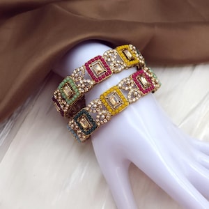 May include: Two ornate bangles with square, multi-colored gemstone settings. The bangles feature a gold-toned metal base with alternating squares of red, yellow, green, and blue gemstones. The design includes small, round, clear stones between the squares.