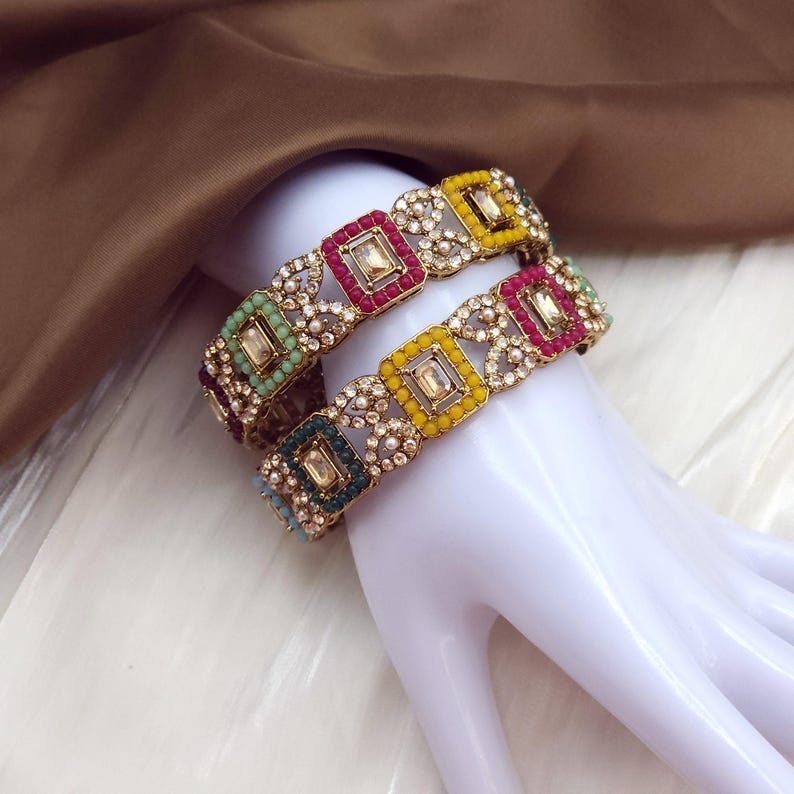 May include: Two ornate bangles with square, multi-colored gemstone settings. The bangles feature a gold-toned metal base with alternating squares of red, yellow, green, and blue gemstones. The design includes small, round, clear stones between the squares.