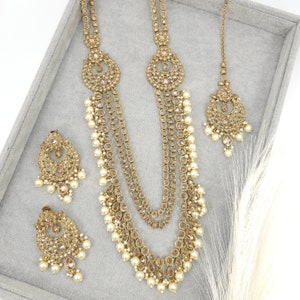Op de afbeelding: Een goudkleurige sieradenset met een lange ketting, oorbellen en een hoofdband. De ketting heeft meerdere strengen met parelaccenten. De oorbellen en hoofdband weerspiegelen het ontwerp van de ketting, wat zorgt voor een samenhangende en elegante uitstraling. De set wordt gepresenteerd op een grijze ondergrond.