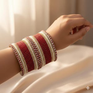 May include: A collection of red, white, and gold bangles encircles a wrist. The bangles showcase alternating bands of red, small white beads, and gold detailing. The jewellery is presented against a soft, neutral backdrop.