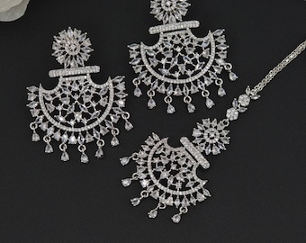 May include: A set of silver earrings and a matching necklace with clear crystal embellishments. The earrings feature a crescent moon design with dangling crystals. The necklace has a similar design with a smaller crescent moon pendant.