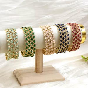 May include: Five gold-coloured bangles, each set with teardrop-shaped gemstones in light green, emerald green, pale pink, navy blue, and ruby red. The bangles are displayed on a white cylindrical stand with a wooden base, showcasing the jewellery.