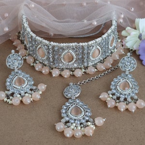 Peut inclure: Un ensemble de bijoux de mari&eacute;e argent&eacute;s comprenant un collier ras du cou, des boucles d'oreilles et un bijou de t&ecirc;te. L'ensemble comprend des pierres en forme de goutte d'eau de couleur p&ecirc;che, de petites perles blanches et des perles pendantes de couleur p&ecirc;che. Les bijoux sont pr&eacute;sent&eacute;s sur un tissu rose doux.