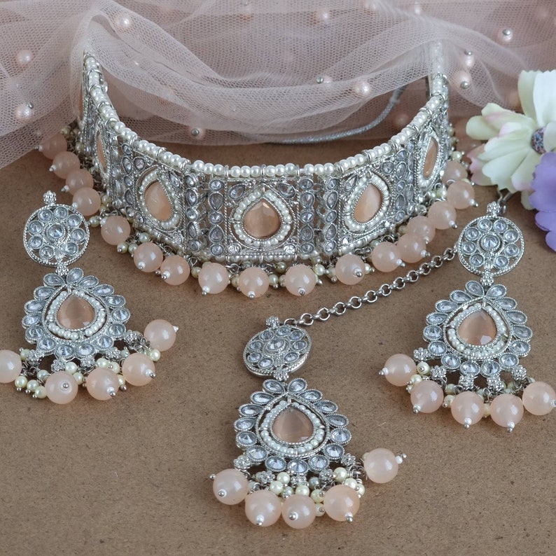 Peut inclure: Un ensemble de bijoux de mari&eacute;e argent&eacute;s comprenant un collier ras du cou, des boucles d'oreilles et un bijou de t&ecirc;te. L'ensemble comprend des pierres en forme de goutte d'eau de couleur p&ecirc;che, de petites perles blanches et des perles pendantes de couleur p&ecirc;che. Les bijoux sont pr&eacute;sent&eacute;s sur un tissu rose doux.