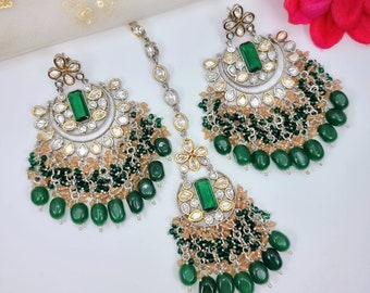 May include: A set of silver and gold earrings and a matching maang tikka. The earrings and tikka feature a crescent moon design with green and clear stones. The earrings have a fringe of green and beige beads.