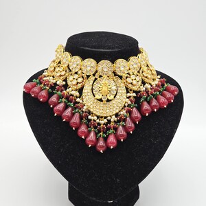 Pu&ograve; includere: Un collier choker dorato con un design a forma di mezzaluna e accenti di pietre preziose rosse e verdi. Il collier &egrave; su un supporto in velluto nero.