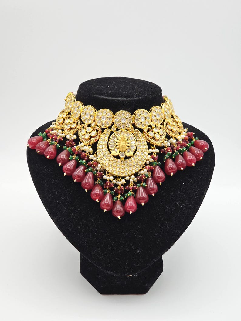 Pu&ograve; includere: Un collier choker dorato con un design a forma di mezzaluna e accenti di pietre preziose rosse e verdi. Il collier &egrave; su un supporto in velluto nero.