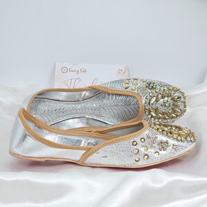 Peut inclure: Une paire de chaussures plates argent&eacute;es et dor&eacute;es orn&eacute;es d'un liser&eacute; en cuir beige. Les chaussures ont une pointe pointue et sont d&eacute;cor&eacute;es de strass et de perles.