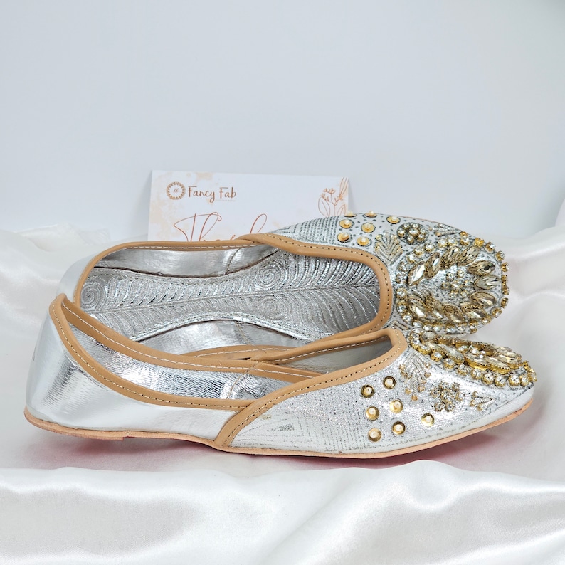 Peut inclure: Une paire de chaussures plates argent&eacute;es et dor&eacute;es orn&eacute;es d'un liser&eacute; en cuir beige. Les chaussures ont une pointe pointue et sont d&eacute;cor&eacute;es de strass et de perles.
