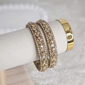 May include: Two gold-toned bangles with intricate geometric patterns and clear stone accents. A plain gold bangle is also visible. The jewelry is displayed on a white cylindrical stand, suggesting a focus on accessories.
