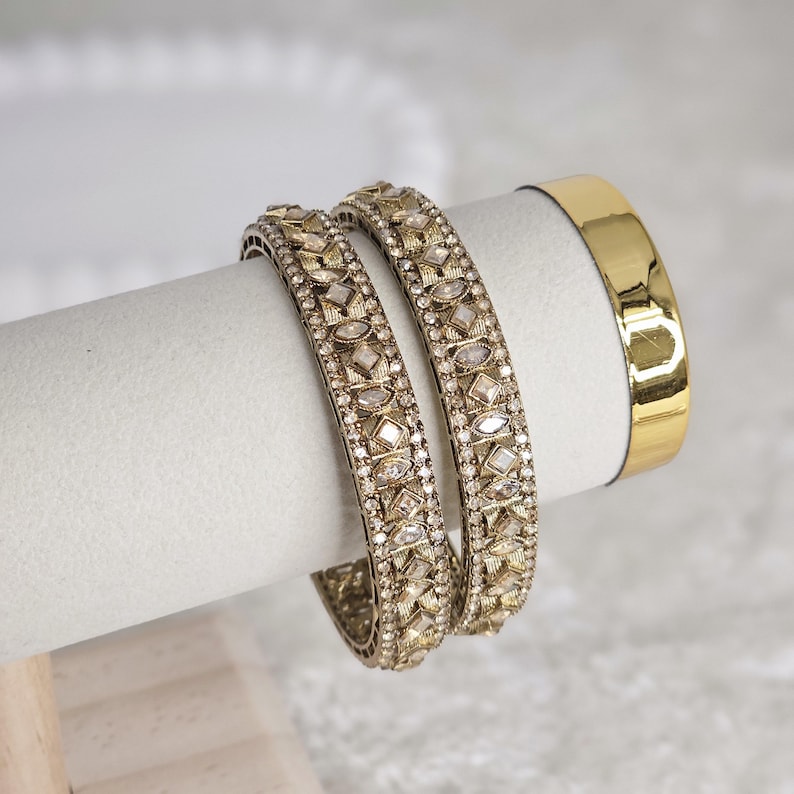 May include: Two gold-toned bangles with intricate geometric patterns and clear stone accents. A plain gold bangle is also visible. The jewelry is displayed on a white cylindrical stand, suggesting a focus on accessories.