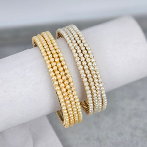 May include: Two sets of pearl bracelets, one set is a light cream colour and the other is a white colour. Both sets are made of small, round pearls and are stacked on top of each other.
