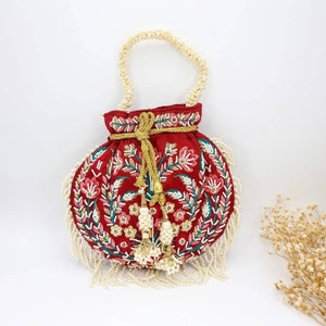 May include: A red embroidered drawstring bag with a pearl handle and tassel accents. The bag is decorated with floral patterns in shades of pink, green, and white.