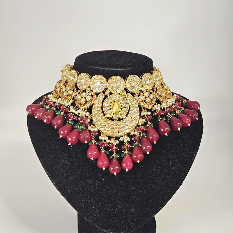 Pu&ograve; includere: Una collana in oro e argento con accenti di pietre preziose rosse e verdi. La collana presenta un grande ciondolo decorato con un motivo floreale. Il ciondolo &egrave; circondato da pietre preziose rotonde pi&ugrave; piccole.