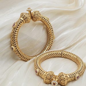 Peut inclure: Deux bracelets dorés ornés de motifs complexes et de petites pierres rouges. Les bracelets sont présentés sur une surface blanche et texturée. Le design comprend des filigranes et des fermoirs décoratifs.