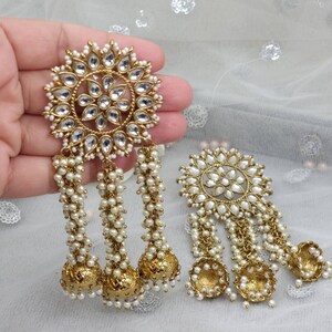 May include: A pair of gold earrings with white pearl accents. The earrings feature a large round design with a floral pattern and three dangling strands of pearls. Each strand ends with a gold bell.