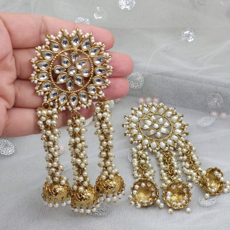 May include: A pair of gold earrings with white pearl accents. The earrings feature a large round design with a floral pattern and three dangling strands of pearls. Each strand ends with a gold bell.