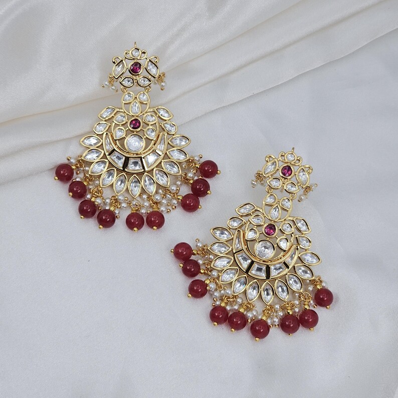 May include: Gold-toned earrings with a floral and crescent moon design. The earrings feature clear and ruby-colored stones, with small white pearls and ruby-colored beads. The earrings are displayed on a white, textured surface.