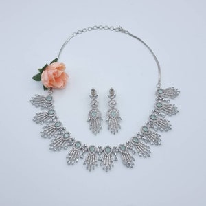 May include: A silver-toned jewelry set featuring a necklace and matching earrings. The necklace has a series of decorative elements with clear and light green accents. The earrings have a similar design. A peach-colored flower is placed near the jewelry.