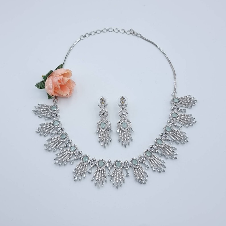 May include: A silver-toned jewelry set featuring a necklace and matching earrings. The necklace has a series of decorative elements with clear and light green accents. The earrings have a similar design. A peach-colored flower is placed near the jewelry.