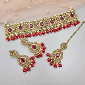 Peut inclure: Un ensemble de bijoux de couleur or antique comprenant un collier ras du cou, des boucles d'oreilles et un serre-t&ecirc;te. L'ensemble est orn&eacute; de pierres pr&eacute;cieuses rouges en forme de goutte, de petites perles blanches et d'accents de perles rouges. Le collier et les boucles d'oreilles ont un design assorti.
