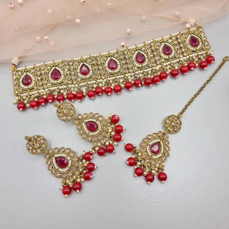Peut inclure: Un ensemble de bijoux de couleur or antique comprenant un collier ras du cou, des boucles d'oreilles et un serre-t&ecirc;te. L'ensemble est orn&eacute; de pierres pr&eacute;cieuses rouges en forme de goutte, de petites perles blanches et d'accents de perles rouges. Le collier et les boucles d'oreilles ont un design assorti.