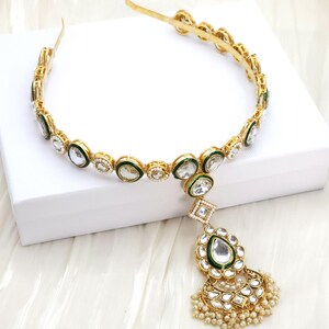 May include: Gold-toned headband with clear and green accents. The headband features a central pendant with a large clear stone, green border, and pearl-like beads. The design is ornate and elegant.