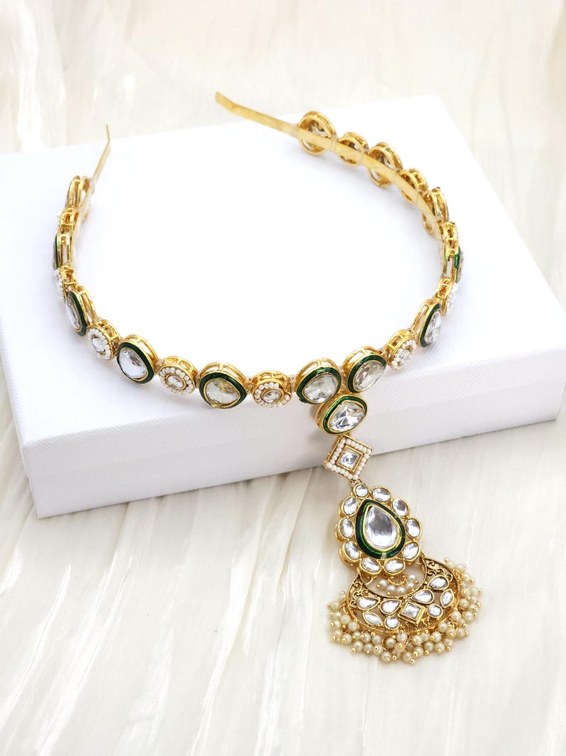 May include: Gold-toned headband with clear and green accents. The headband features a central pendant with a large clear stone, green border, and pearl-like beads. The design is ornate and elegant.