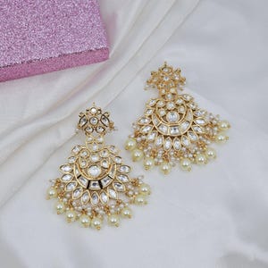 May include: A pair of gold-toned chandelier earrings with clear, sparkling stones and dangling faux pearls. The earrings have a crescent moon shape with intricate detailing and are displayed on a white, silky fabric.