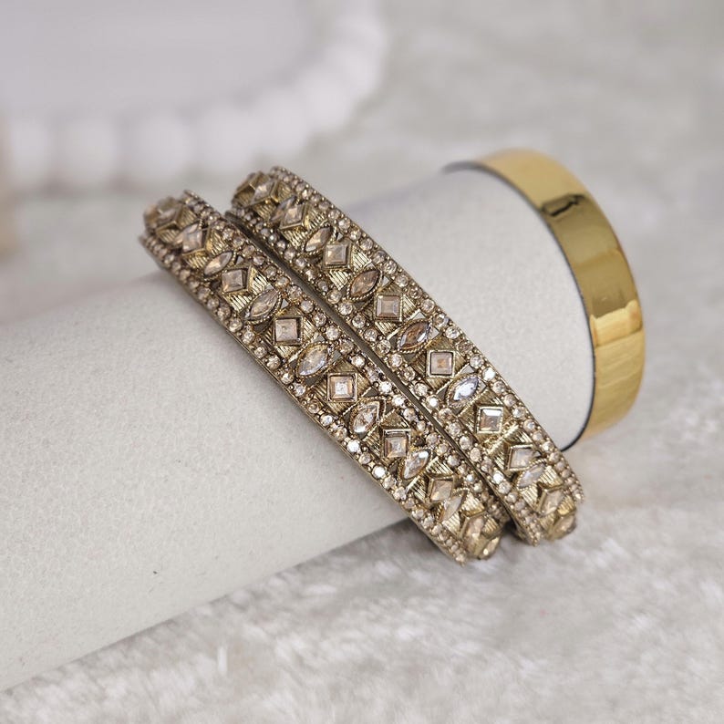 May include: Two ornate gold bangles with clear gemstone accents, featuring a geometric design. A plain gold band is also visible. The jewelry is displayed on a white surface, suggesting an elegant presentation.