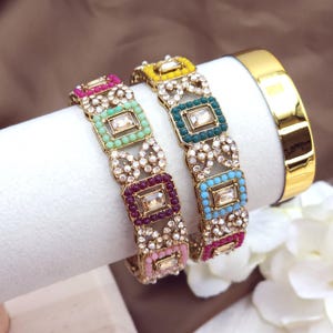 May include: Two ornate gold bangles with colorful square accents and clear rectangular center stones. The bangles feature pink, green, purple, yellow, and blue details, with a gold band on the right.