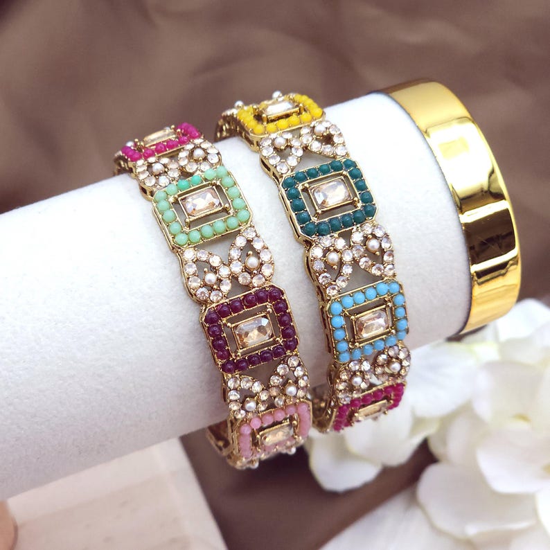 May include: Two ornate gold bangles with colorful square accents and clear rectangular center stones. The bangles feature pink, green, purple, yellow, and blue details, with a gold band on the right.