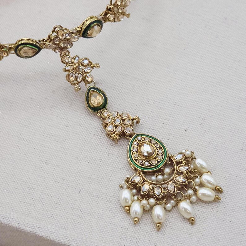 May include: A gold-toned headpiece with intricate detailing. It features clear gemstones, green enamel accents, and dangling faux pearls. The design includes floral and teardrop shapes, creating an elegant and ornate accessory.