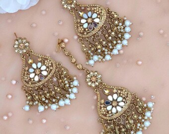 May include: A gold-coloured jewellery set comprising earrings and a headpiece. The earrings and headpiece feature a floral design with white and iridescent accents, and dangling white pearl beads. The set is displayed on a pink surface.