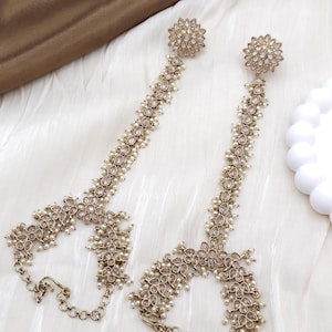 May include: A pair of ornate, gold-toned hand chain jewelry. Each piece features a floral stud, a chain of small, clear stones, and a chain of small pearls. The design includes a chain that connects to a ring. The jewelry is displayed on a white, textured surface.