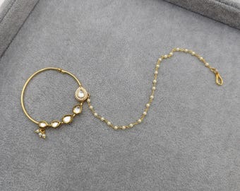 Op de afbeelding: Een gouden neusring met een ketting en witte parel accenten. De ring is rond en heeft een traanvormige hanger met een witte steen.