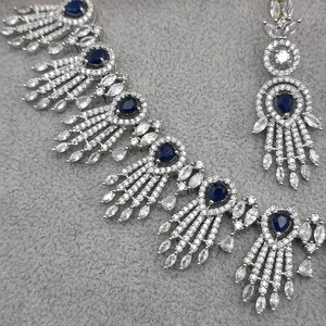 May include: A silver-toned jewelry set featuring a necklace and earrings. The necklace has a series of navy blue gemstone accents surrounded by clear, sparkling stones. The earrings mirror the necklace design, with a navy blue center stone and dangling clear stones.