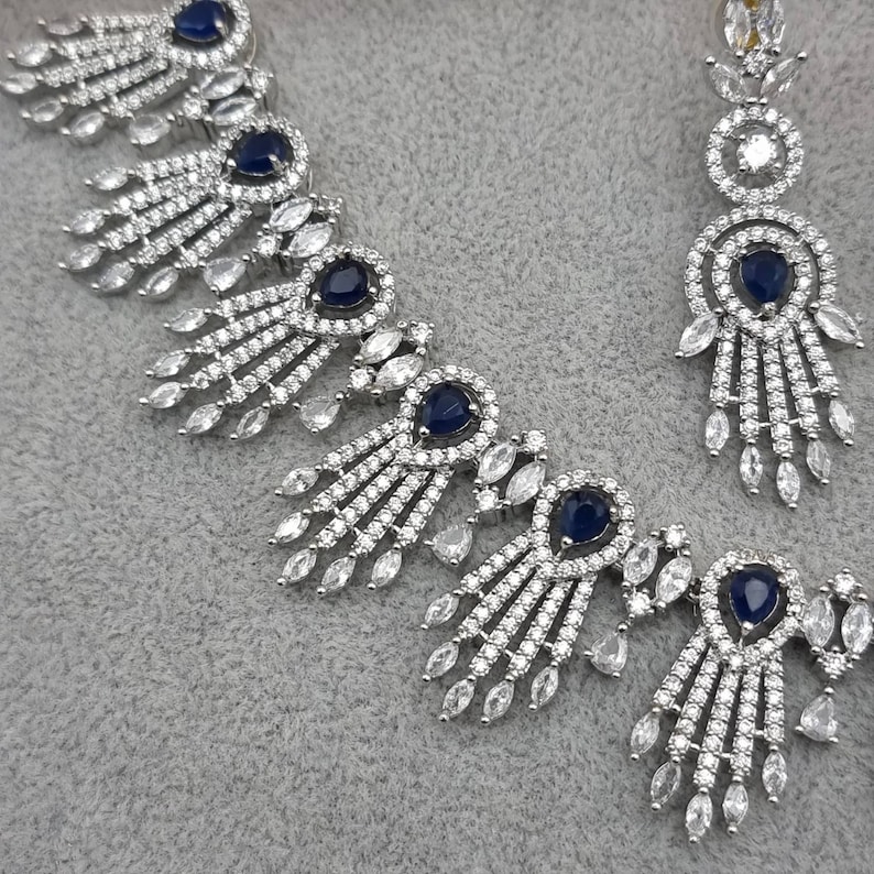 May include: A silver-toned jewelry set featuring a necklace and earrings. The necklace has a series of navy blue gemstone accents surrounded by clear, sparkling stones. The earrings mirror the necklace design, with a navy blue center stone and dangling clear stones.