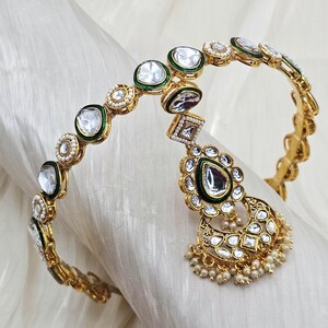 May include: Gold-toned headpiece with clear, round, and square-cut stones, accented with green enamel and small pearls. A teardrop-shaped pendant with a large stone and pearl fringe hangs from the center. The headpiece is displayed on a white fabric.