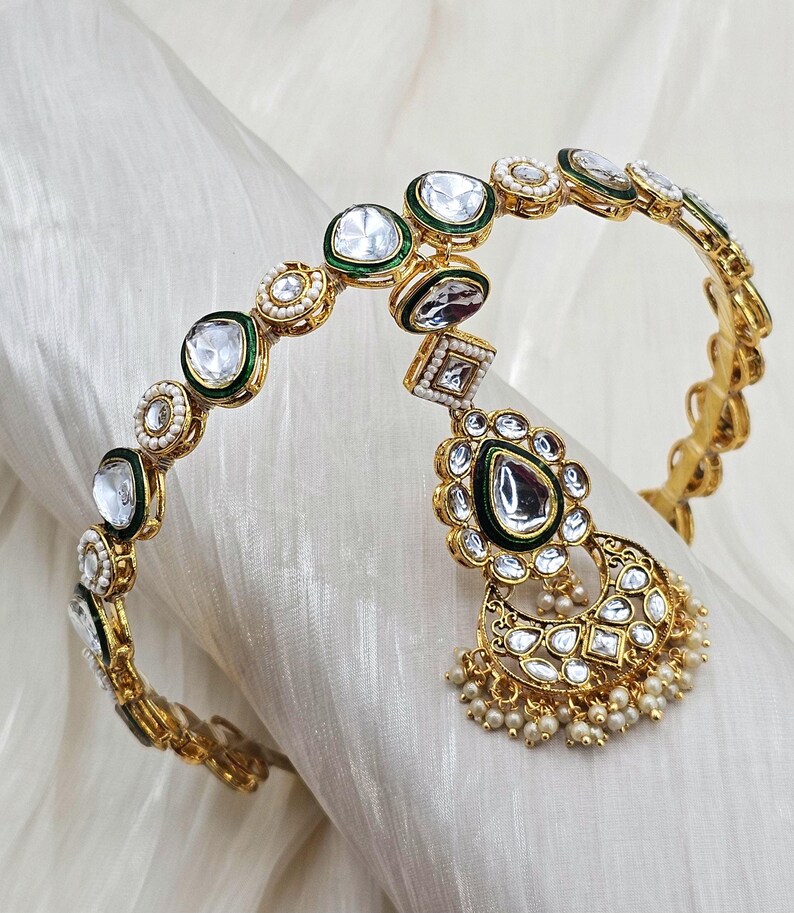 May include: Gold-toned headpiece with clear, round, and square-cut stones, accented with green enamel and small pearls. A teardrop-shaped pendant with a large stone and pearl fringe hangs from the center. The headpiece is displayed on a white fabric.