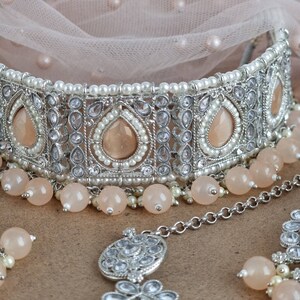 Peut inclure: Un ensemble de bijoux de mari&eacute;e argent&eacute; comprenant un collier ras du cou, un serre-t&ecirc;te et des boucles d'oreilles. Le collier comporte des pierres en forme de goutte de couleur p&ecirc;che, de petites perles et des perles rondes p&ecirc;che. Le serre-t&ecirc;te et les boucles d'oreilles ont des motifs similaires.