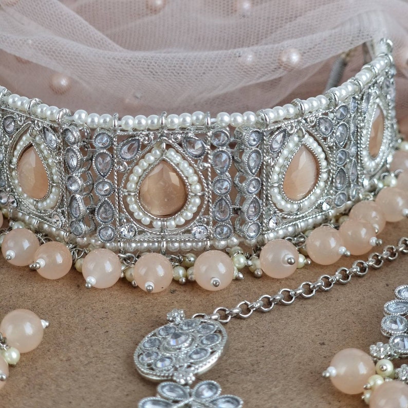 Peut inclure: Un ensemble de bijoux de mari&eacute;e argent&eacute; comprenant un collier ras du cou, un serre-t&ecirc;te et des boucles d'oreilles. Le collier comporte des pierres en forme de goutte de couleur p&ecirc;che, de petites perles et des perles rondes p&ecirc;che. Le serre-t&ecirc;te et les boucles d'oreilles ont des motifs similaires.