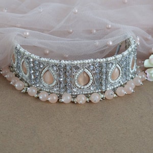 Peut inclure: Un collier ras du cou argent&eacute; orn&eacute; de pierres en forme de goutte de couleur p&ecirc;che et de petites perles blanches. Le collier est orn&eacute; d'un rang de perles rondes p&ecirc;che. Fond en tissu rose clair.