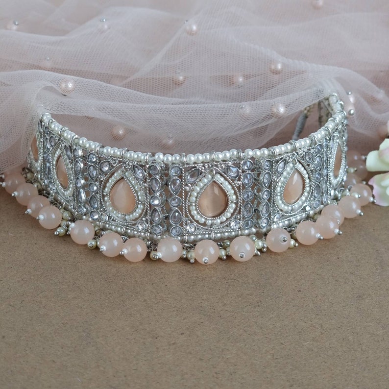 Peut inclure: Un collier ras du cou argent&eacute; orn&eacute; de pierres en forme de goutte de couleur p&ecirc;che et de petites perles blanches. Le collier est orn&eacute; d'un rang de perles rondes p&ecirc;che. Fond en tissu rose clair.