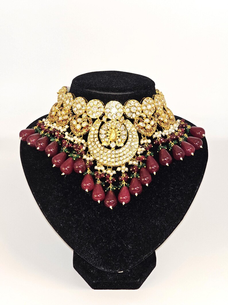 Pu&ograve; includere: Una collana in oro e argento con un grande ciondolo decorato e una frangia di perline rosse e verdi.