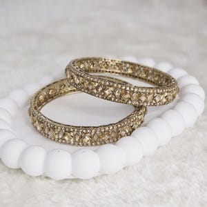 May include: Two gold-toned bangles with intricate geometric designs and embedded gemstones. The bangles are resting on a white, textured surface, creating a contrast between the warm metal and the cool background.